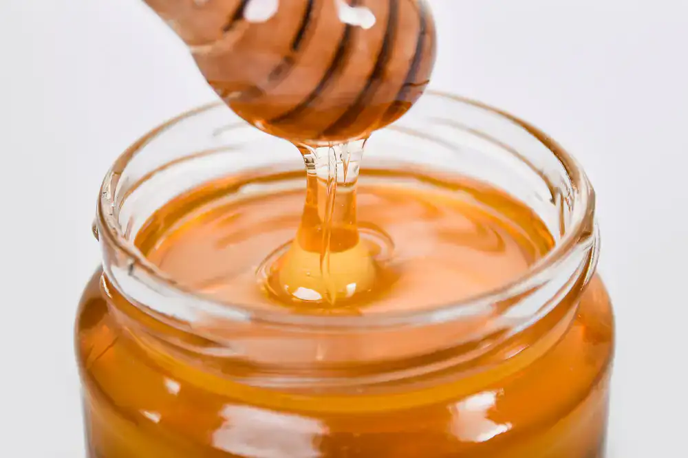 Golden honey pouring smoothly from a wooden dipper into a glass jar